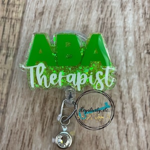 May include: Green glitter badge reel with the text "ABA Therapist" in white lettering. The reel has a clear plastic retractable cord and a clear plastic charm with a crystal rhinestone.