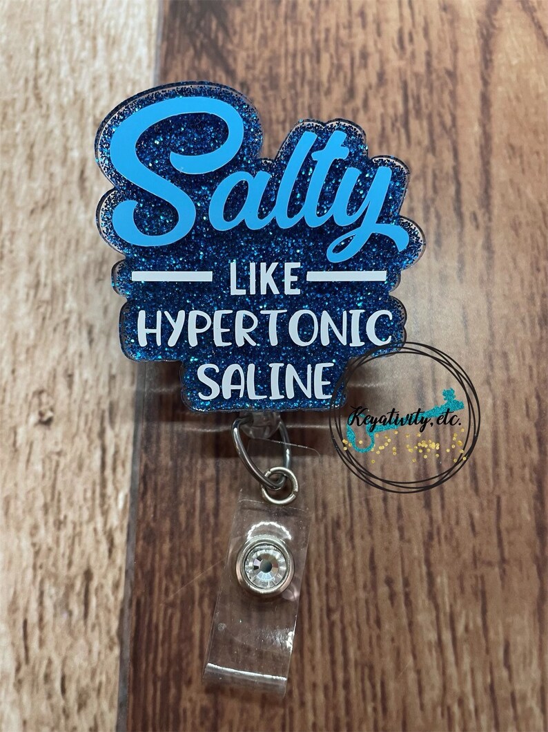 K&ouml;nnte beinhalten: Ein blau glitzernder Ausweis-Clip mit den Worten "Salty like Hypertonic Saline" in wei&szlig;er und blauer Schrift. Der Ausweis-Clip hat einen transparenten Kunststoffclip und einen dekorativen Schmuckstein.