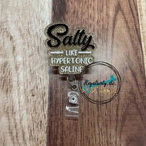 K&ouml;nnte beinhalten: Ein glitzerndes Ausweis-Jojo mit den Worten "Salty like Hypertonic Saline" in schwarzer Schrift und goldfarbenen Buchstaben. Das Ausweis-Jojo hat einen transparenten Kunststoffriemen und einen silbernen Clip. Der Hintergrund ist eine Holzoberfl&auml;che.