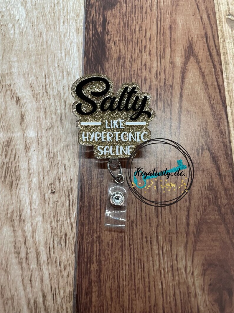 K&ouml;nnte beinhalten: Ein glitzerndes Ausweis-Jojo mit den Worten "Salty like Hypertonic Saline" in schwarzer Schrift und goldfarbenen Buchstaben. Das Ausweis-Jojo hat einen transparenten Kunststoffriemen und einen silbernen Clip. Der Hintergrund ist eine Holzoberfl&auml;che.