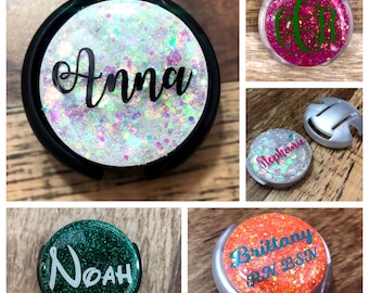 May include: Four round, personalized, glitter-covered, badge holders with a silver metal clip. The names on the badges are Anna, Noah, Brittany RN BSN, and Stephanie.