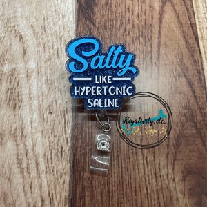 K&ouml;nnte beinhalten: Ein blau glitzernder Ausweis-Rollenhalter mit dem Text "Salty like hypertonic saline". Der Ausweis-Rollenhalter hat einen transparenten Kunststoffclip und eine einziehbare Kordel. Der Hintergrund ist eine Holzoberfl&auml;che.