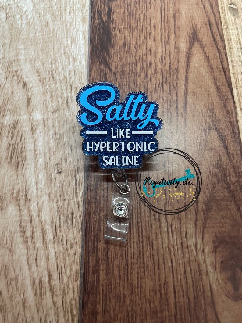 K&ouml;nnte beinhalten: Ein blau glitzernder Ausweis-Rollenhalter mit dem Text "Salty like hypertonic saline". Der Ausweis-Rollenhalter hat einen transparenten Kunststoffclip und eine einziehbare Kordel. Der Hintergrund ist eine Holzoberfl&auml;che.