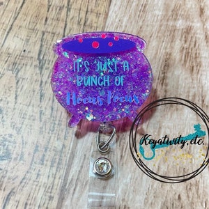 May include: A purple glitter badge reel in the shape of a cauldron, with the text "It's Just a Bunch of Hocus Pocus". The cauldron has a purple rim and is decorated with red dots. The badge reel has a clear plastic clip and a silver metal clip.
