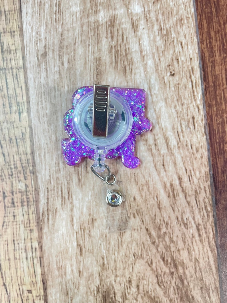 Op de afbeelding: Paarse glitter badge reel met een transparante plastic badgehouder en een zilveren clip. De reel heeft een transparante plastic bedel met een zilveren strass.