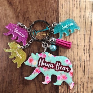 May include: A collection of bear-shaped keychains in various colors, including pink, yellow, and teal. Each keychain features a name, with one reading "Nana Bear" in a floral design. The keychains are attached to a silver keyring with a pink tassel.