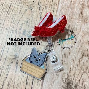 May include: A badge reel featuring a pair of sparkling red ruby slippers and a dog in a basket charm. The reel has a clear plastic strap and a decorative crystal. The text "*BADGE REEL NOT INCLUDED" is visible.