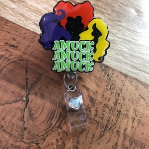 May include: A badge reel featuring the silhouettes of three witches' hair in purple, red, and yellow, with the text "AMUCK AMUCK AMUCK" in green. The badge reel has a clear plastic strap and a silver-colored clip.