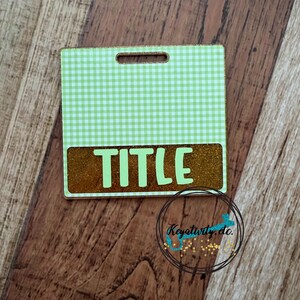May include: A rectangular badge with a green gingham pattern top and a gold glitter bottom. The word "TITLE" is in large, light green letters. The badge has a small hole at the top for attachment.