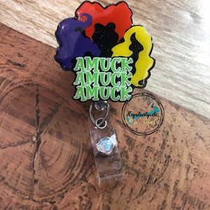 May include: A badge reel featuring the silhouettes of three witches' hair in red, yellow, and purple, with the text "AMUCK AMUCK AMUCK" in green. The reel has a clear plastic strap and a silver-colored clip.