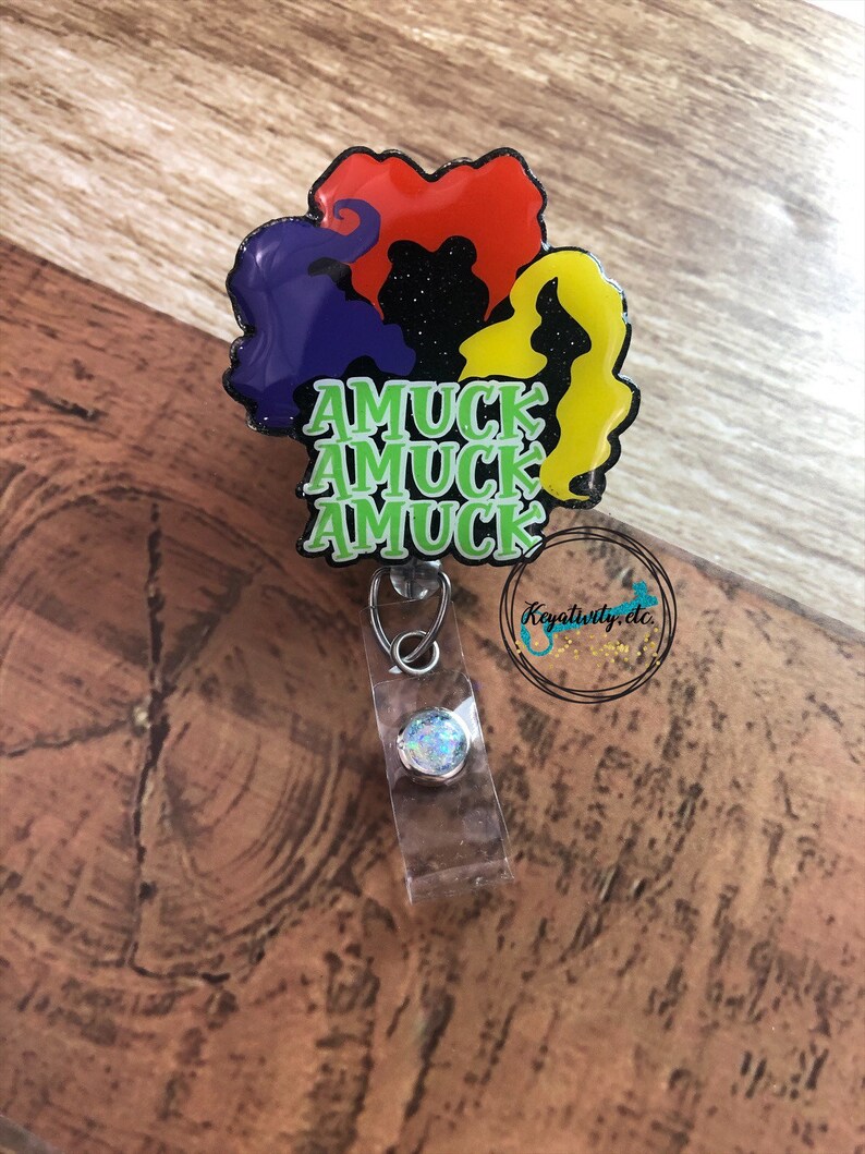 May include: A badge reel featuring the silhouettes of three witches' hair in red, yellow, and purple, with the text "AMUCK AMUCK AMUCK" in green. The reel has a clear plastic strap and a silver-colored clip.