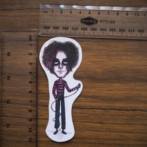 May include: A sticker featuring a cartoon illustration of a person with dark hair and a pink and black striped shirt holding a microphone. The person is wearing black pants and black shoes.