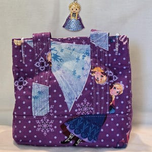May include: A small, purple tote bag with a repeating pattern of white polka dots and snowflakes, featuring images of a cartoon character. A beaded necklace with a pendant of the same character hangs above the bag. The necklace has purple and blue beads.