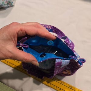 May include: A small tote bag with a purple floral pattern and a bright blue interior. The bag has two small handles and a magnetic snap closure. The bag is held open, revealing the interior.