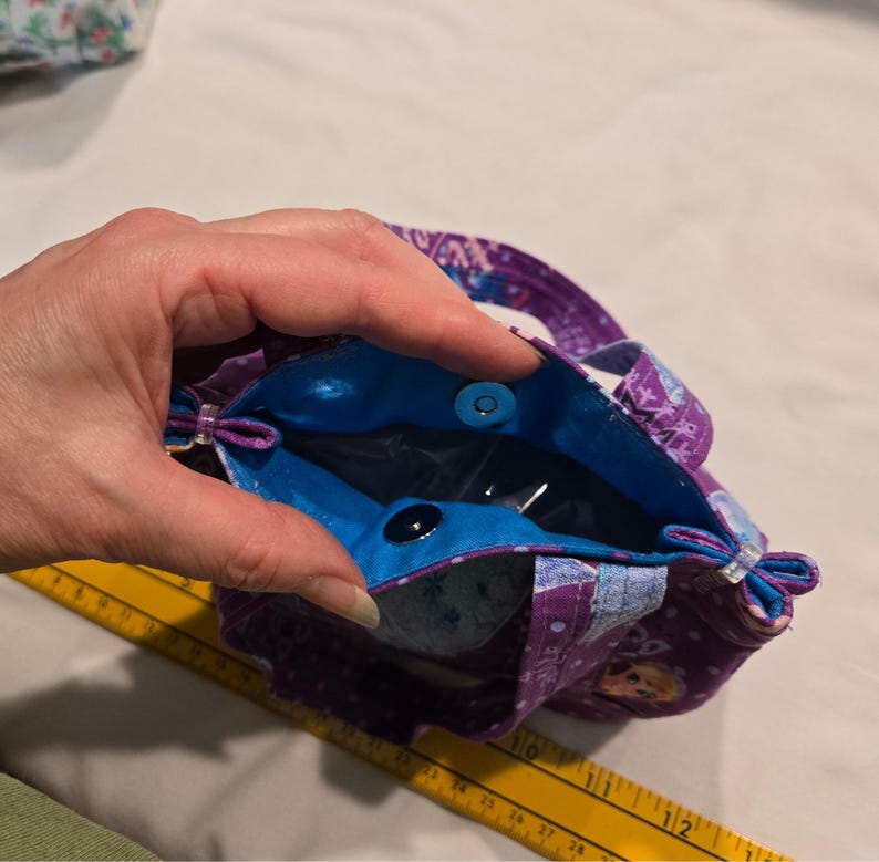 May include: A small tote bag with a purple floral pattern and a bright blue interior. The bag has two small handles and a magnetic snap closure. The bag is held open, revealing the interior.