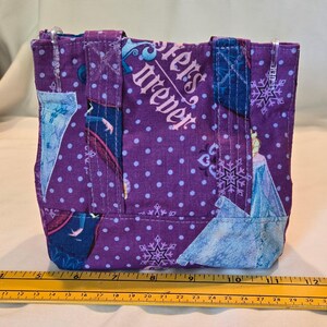 May include: A small, purple tote bag with a repeating pattern of white polka dots and snowflakes. The bag features images of a princess and the text "Sisters Forever". The bag has two handles and is approximately 20 cm wide.