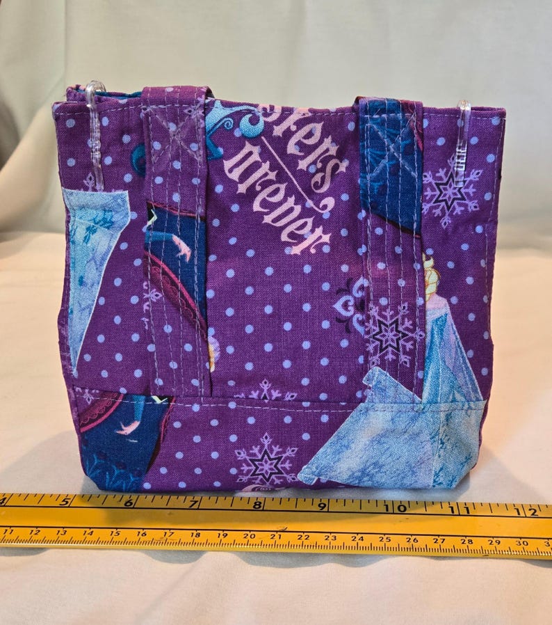 May include: A small, purple tote bag with a repeating pattern of white polka dots and snowflakes. The bag features images of a princess and the text "Sisters Forever". The bag has two handles and is approximately 20 cm wide.
