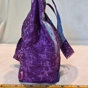 May include: A handmade fabric tote bag in shades of purple, blue, and pink. The bag features a textured, patterned design and sturdy handles. The bag's base measures approximately 15 cm.