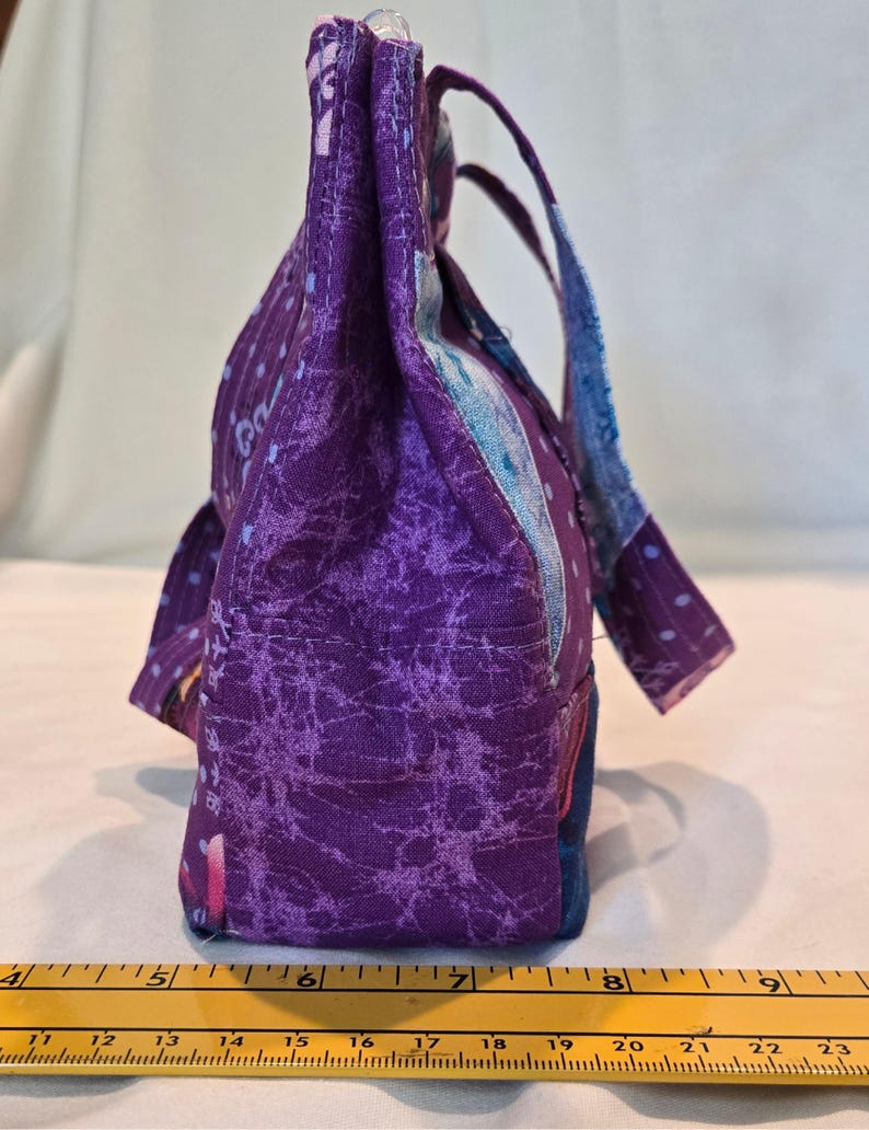 May include: A handmade fabric tote bag in shades of purple, blue, and pink. The bag features a textured, patterned design and sturdy handles. The bag's base measures approximately 15 cm.