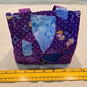 May include: A small, handmade tote bag featuring a purple fabric with white polka dots and snowflake patterns. The bag has two handles and patches of blue fabric. The design includes images of a cartoon character. The bag measures approximately 13 cm wide.
