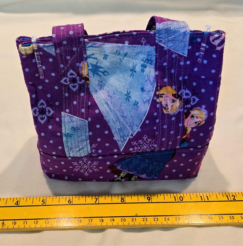 May include: A small, handmade tote bag featuring a purple fabric with white polka dots and snowflake patterns. The bag has two handles and patches of blue fabric. The design includes images of a cartoon character. The bag measures approximately 13 cm wide.