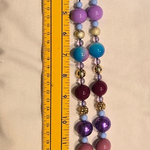 May include: A beaded necklace with a blue ribbon, featuring various sized beads in shades of purple, blue, and gold. The necklace is approximately 40 cm long and includes a charm of a doll.