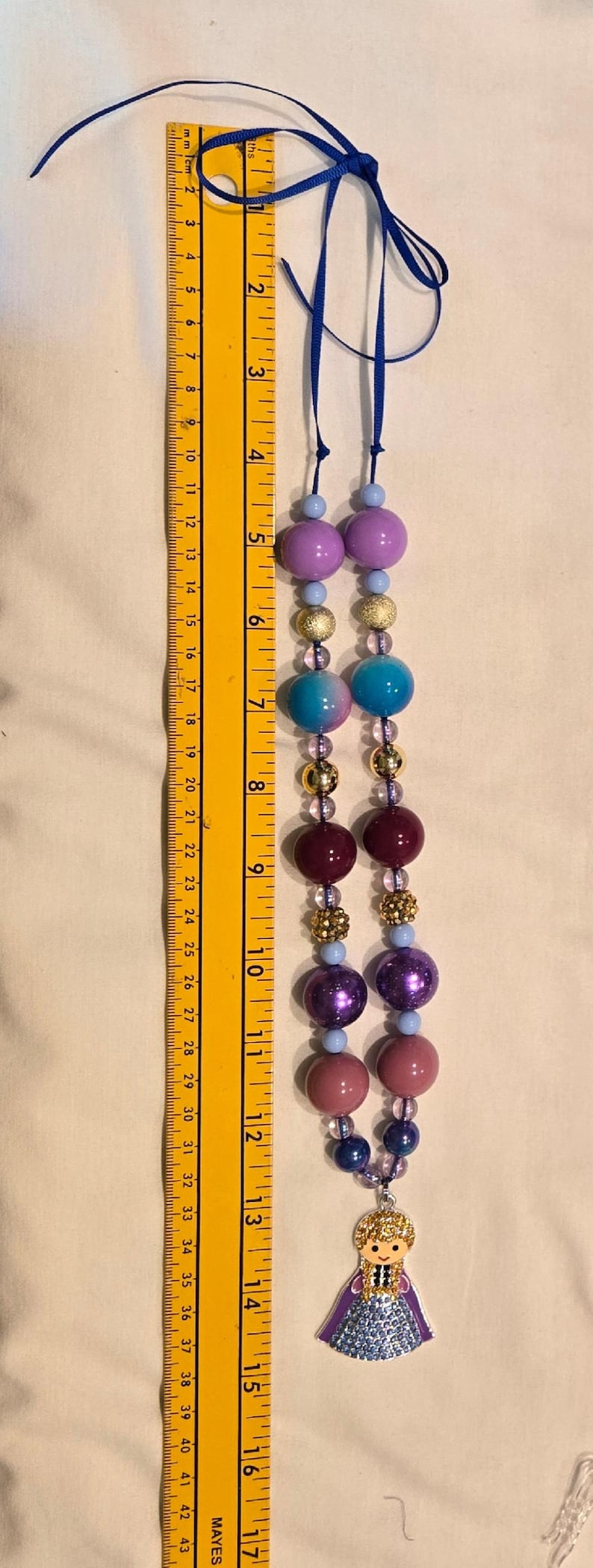 May include: A beaded necklace with a blue ribbon, featuring various sized beads in shades of purple, blue, and gold. The necklace is approximately 40 cm long and includes a charm of a doll.