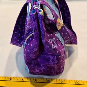May include: A handmade purple fabric handbag with a patterned design. The bag features a top handle and a shoulder strap. The fabric has a marbled effect and is accented with white polka dots. The bag is approximately 25 cm wide.