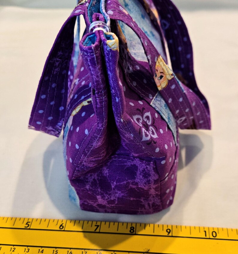 May include: A handmade purple fabric handbag with a patterned design. The bag features a top handle and a shoulder strap. The fabric has a marbled effect and is accented with white polka dots. The bag is approximately 25 cm wide.