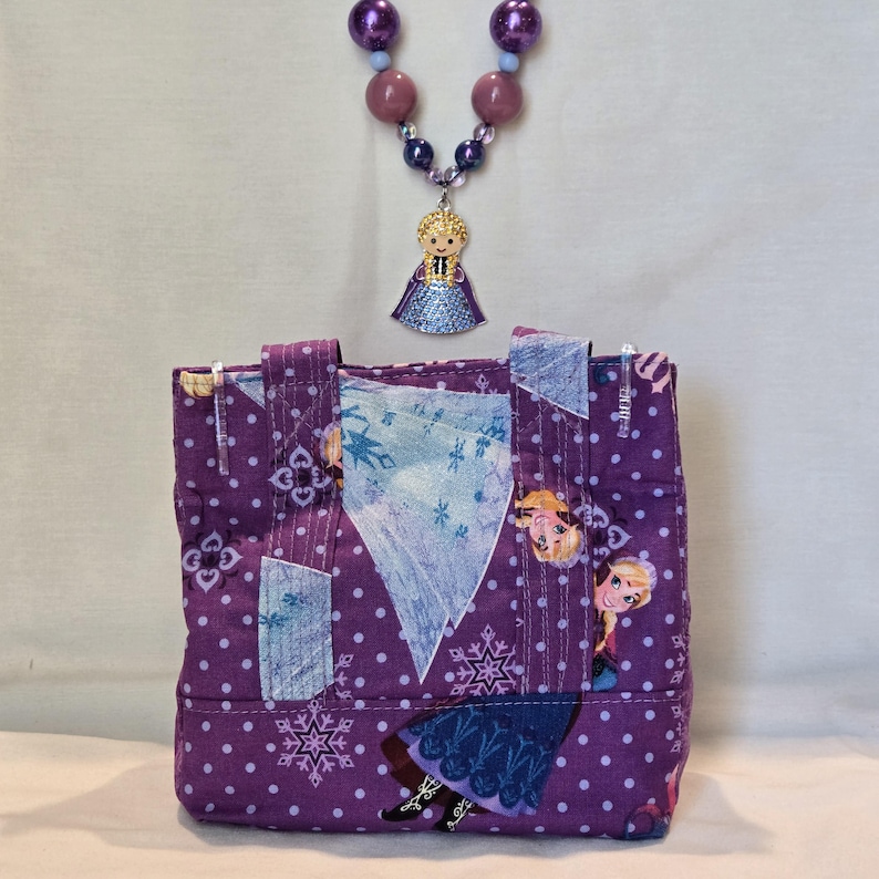 May include: A small, purple tote bag with a repeating pattern of white polka dots and snowflakes, featuring images of a cartoon character. A beaded necklace with a pendant of the same character hangs above the bag. The necklace has purple and blue beads.