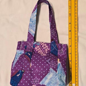 May include: A small, purple tote bag with a polka dot pattern and snowflake and character accents. The bag features the text "Sisters Forever" and has a zipper closure. The bag's handles are approximately 28 cm long.