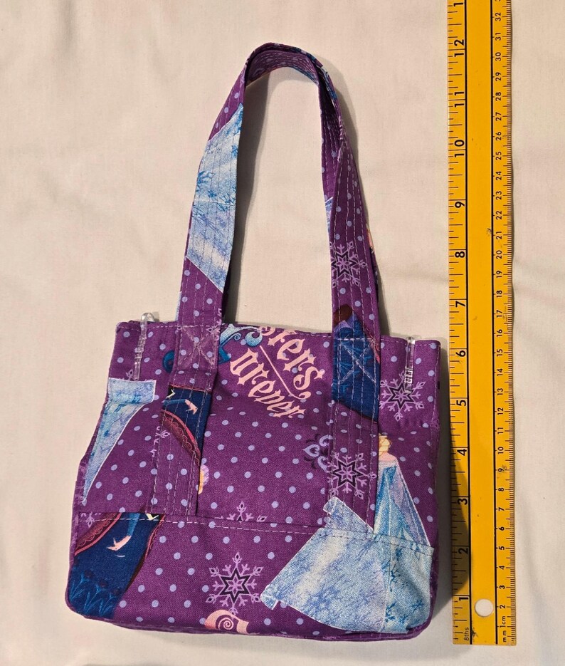 May include: A small, purple tote bag with a polka dot pattern and snowflake and character accents. The bag features the text "Sisters Forever" and has a zipper closure. The bag's handles are approximately 28 cm long.