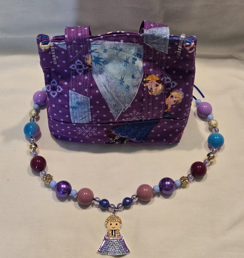 May include: A purple fabric purse with a patchwork design featuring images of Elsa and snowflakes. A beaded necklace with an Elsa pendant is in front of the purse. The necklace has purple, blue, and gold beads.