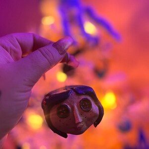 Puede incluir: Un adorno de arcilla hecho a mano de un personaje de la pel&iacute;cula "Coraline" con ojos de bot&oacute;n negros y una cara marr&oacute;n. El adorno cuelga de una mano frente a un fondo borroso de un &aacute;rbol de Navidad con luces blancas.