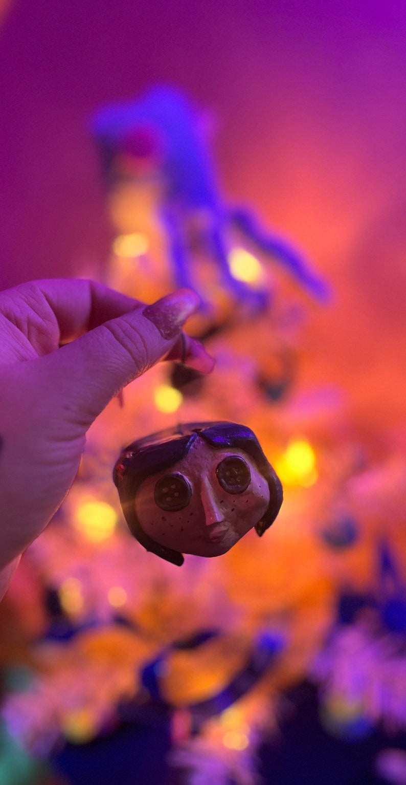 Puede incluir: Un adorno de arcilla hecho a mano de un personaje de la pel&iacute;cula "Coraline" con ojos de bot&oacute;n negros y una cara marr&oacute;n. El adorno cuelga de una mano frente a un fondo borroso de un &aacute;rbol de Navidad con luces blancas.