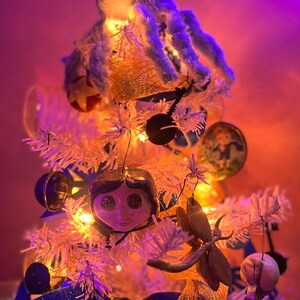 Puede incluir: Un &aacute;rbol de Navidad artificial blanco decorado con un pulpo de peluche azul, un adorno con la cara de una mu&ntilde;eca y una variedad de otros adornos, incluyendo una cinta dorada, una m&aacute;scara azul y dorada y una lib&eacute;lula de madera.