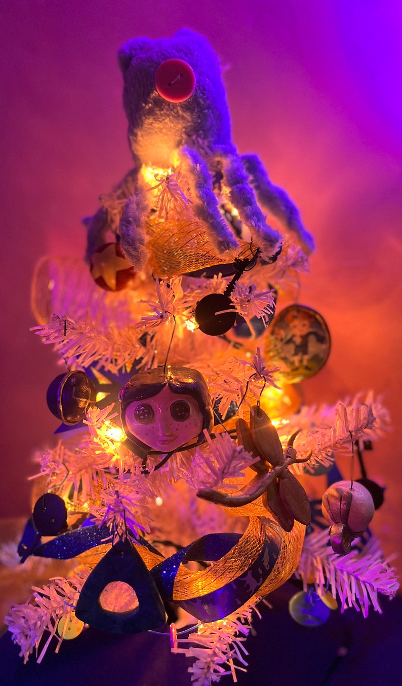 Puede incluir: Un &aacute;rbol de Navidad artificial blanco decorado con un pulpo de peluche azul, un adorno con la cara de una mu&ntilde;eca y una variedad de otros adornos, incluyendo una cinta dorada, una m&aacute;scara azul y dorada y una lib&eacute;lula de madera.