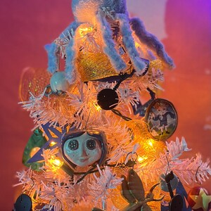 Puede incluir: Un &aacute;rbol de Navidad artificial blanco decorado con un pulpo de peluche azul, una cinta amarilla y azul, una cara de cer&aacute;mica blanca con ojos grandes y otros adornos.
