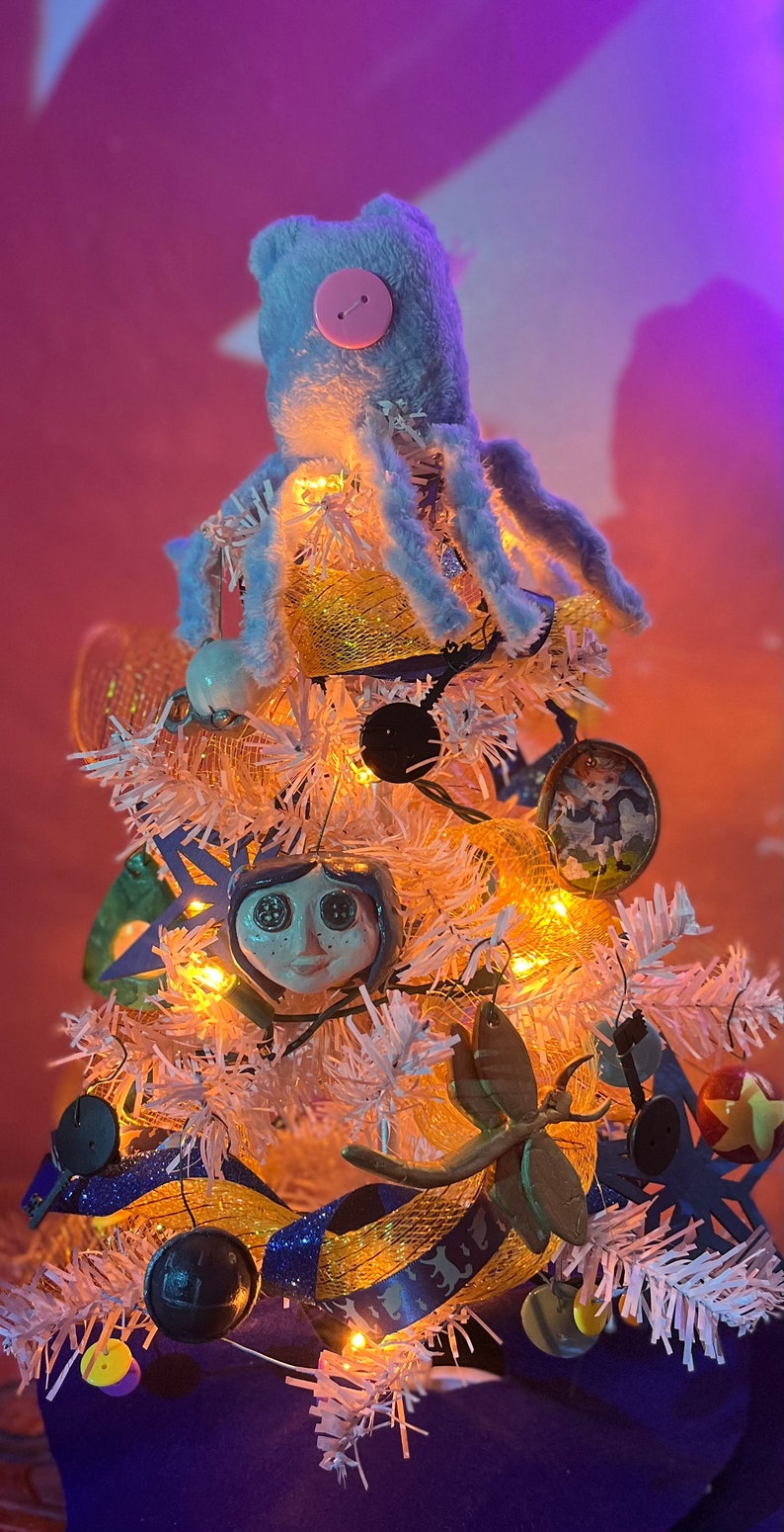 Puede incluir: Un &aacute;rbol de Navidad artificial blanco decorado con un pulpo de peluche azul, una cinta amarilla y azul, una cara de cer&aacute;mica blanca con ojos grandes y otros adornos.
