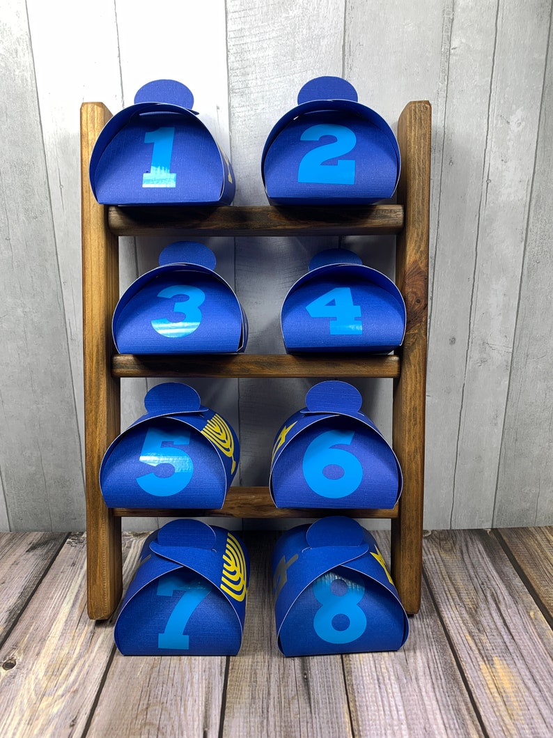 May include: Eight blue gift boxes with numbers 1 through 8 printed on them are displayed on a wooden shelf. Each box has a gold menorah design on the front.