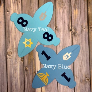 May include: Two blue paper Hanukkah gift boxes with gold accents. The boxes are decorated with a Star of David, a menorah, a dreidel, and the number one and eight. The boxes are labeled "Navy Teal" and "Navy Blue".