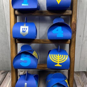 May include: Eight blue gift boxes with different designs for Hanukkah. The boxes are arranged on a wooden shelf. The designs include a Star of David, a menorah, a dreidel, and the numbers 1 through 8.