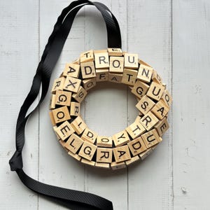 May include: A wooden wreath made of Scrabble tiles spelling out the phrase "Round and Round We Go" with a black ribbon attached.