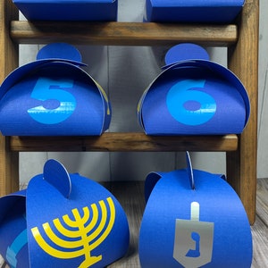 May include: Six blue favor boxes with yellow and silver accents. The boxes are decorated with numbers 3, 4, 5, 6, a menorah, and a dreidel. The boxes are arranged on a wooden shelf.