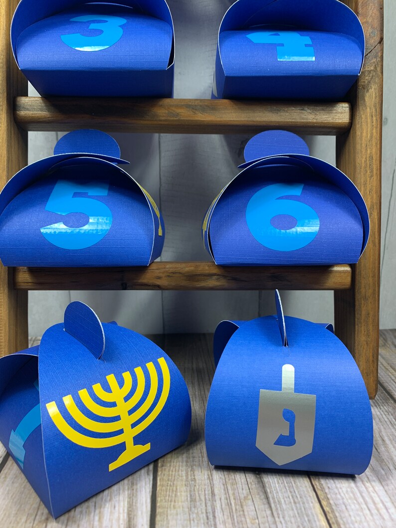 May include: Six blue favor boxes with yellow and silver accents. The boxes are decorated with numbers 3, 4, 5, 6, a menorah, and a dreidel. The boxes are arranged on a wooden shelf.