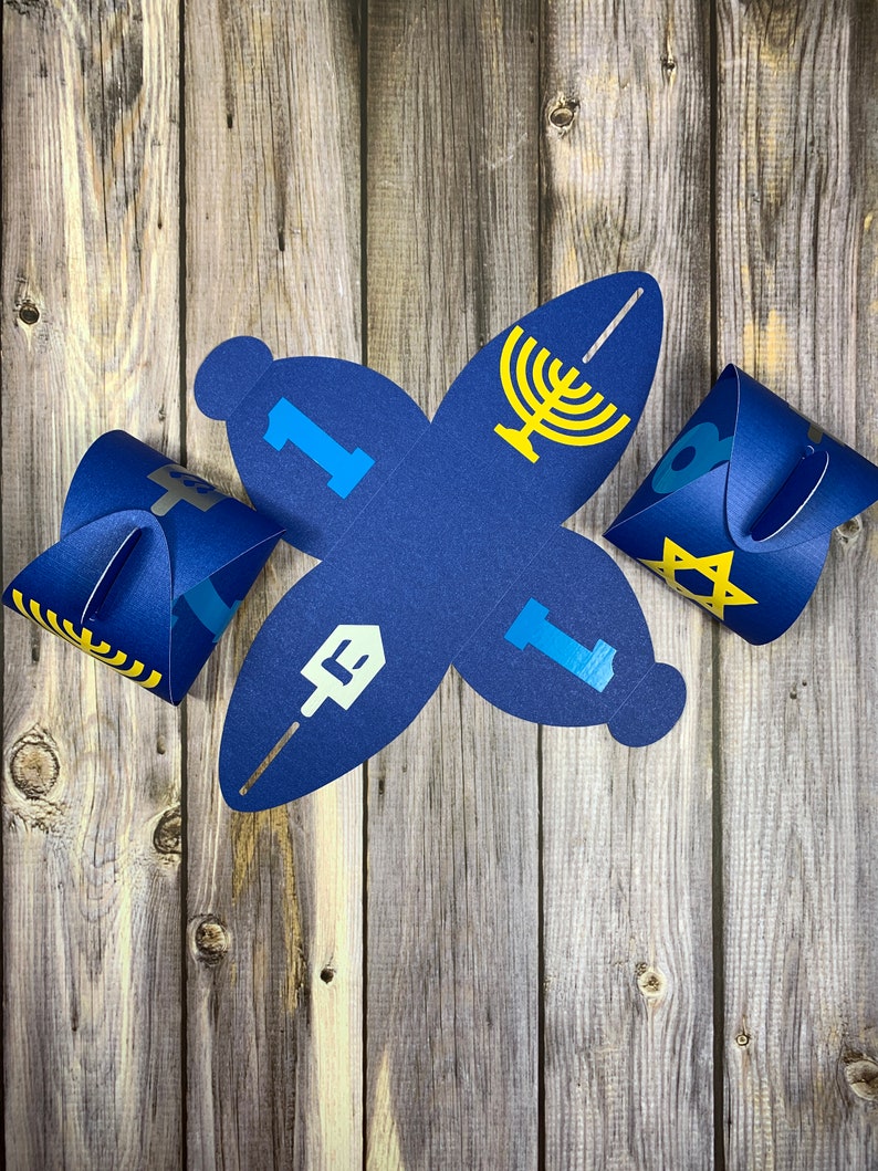 May include: Blue paper gift boxes with yellow and blue designs. The boxes feature symbols associated with Hanukkah, including a menorah, a dreidel, a Star of David, and a Hebrew letter.