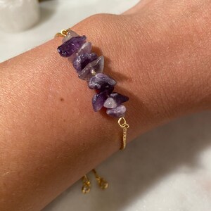 May include: A gold chain bracelet with a row of purple amethyst stones. The bracelet has a sliding clasp.