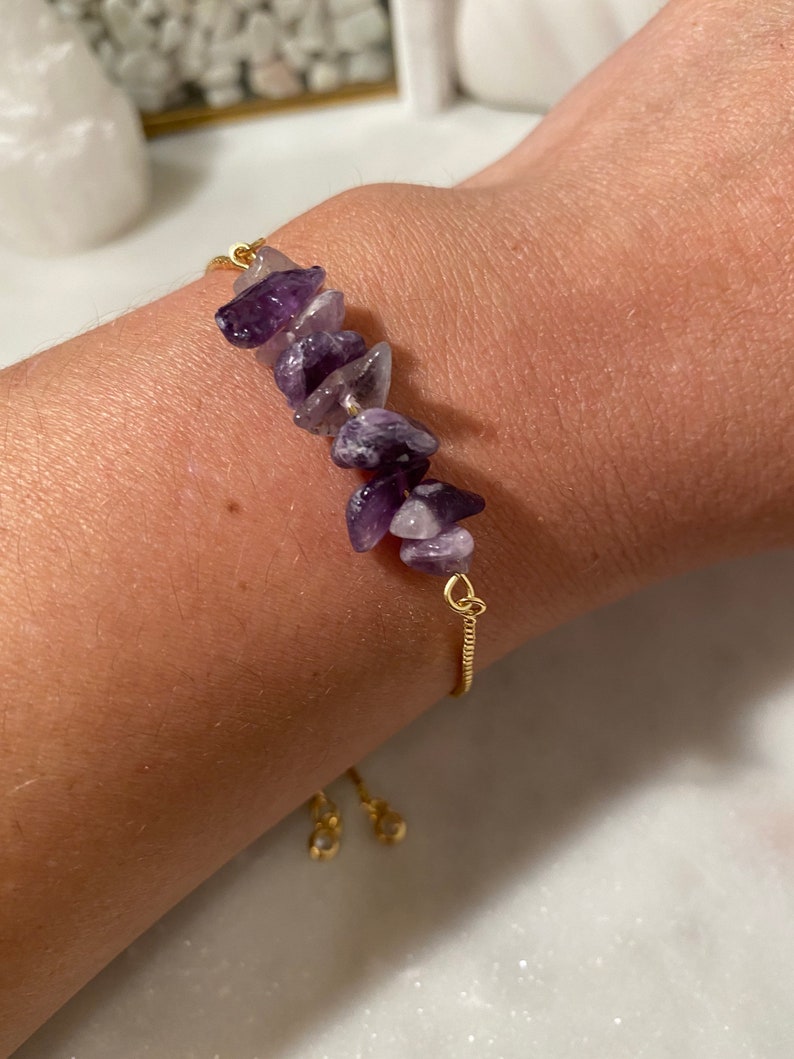 May include: A gold chain bracelet with a row of purple amethyst stones. The bracelet has a sliding clasp.