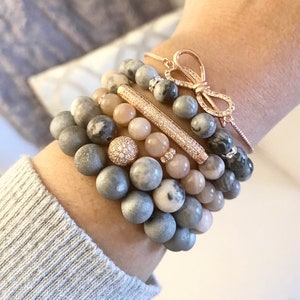 May include: A stack of bracelets with gray and pink stones, a gold bar charm, and a gold bow charm.