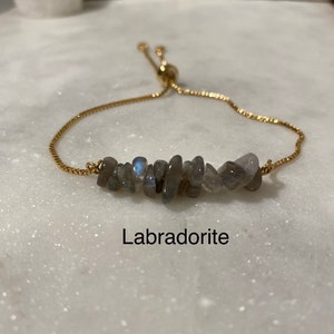 May include: A gold chain bracelet with a slider clasp and a row of grey labradorite stones. The bracelet is on a white surface and the word "Labradorite" is written below the bracelet.